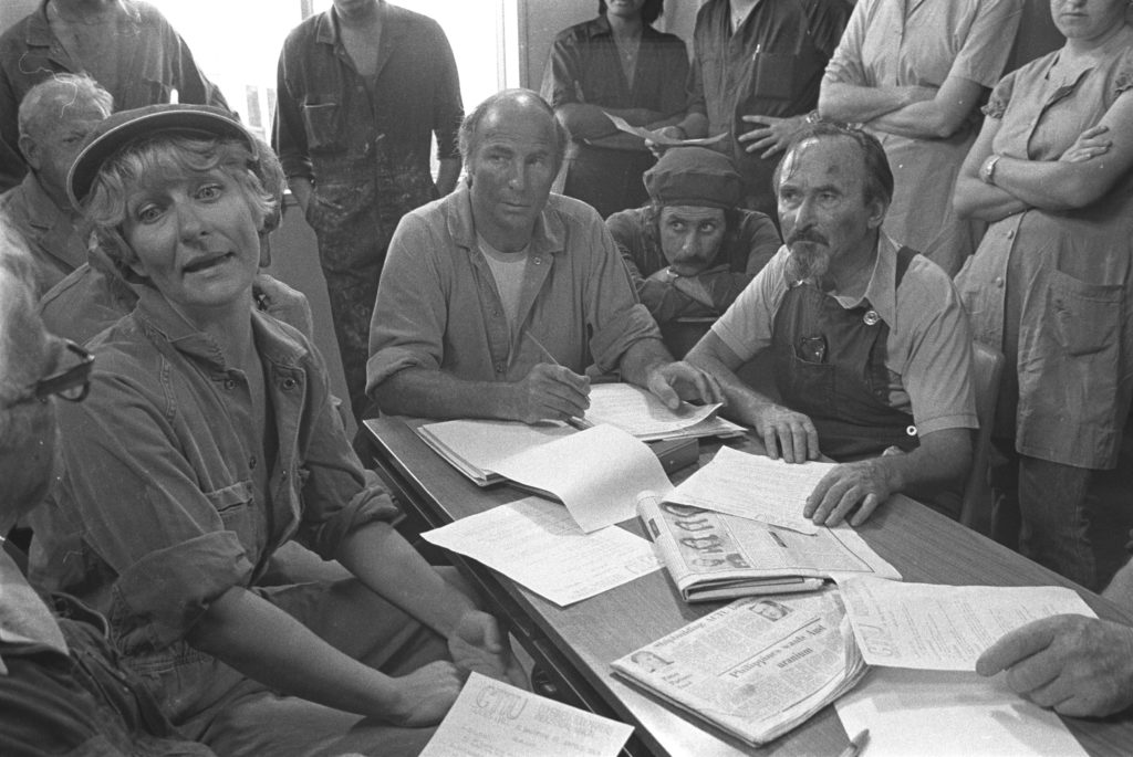 Workers sit around a table with papers discussing the negotiation of a log of claims with their employer. The photograph is in black and white. Four workers are seated, the others stand around in the background.
