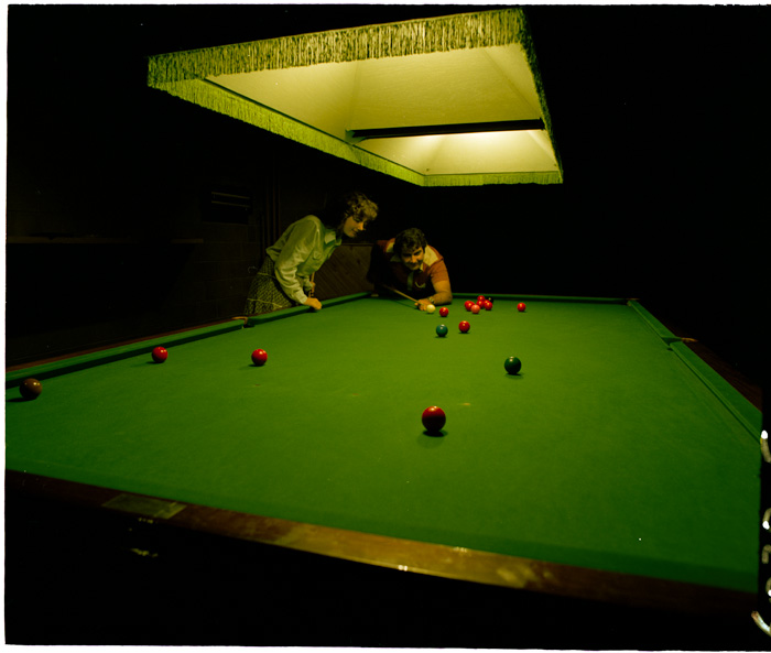 A woman and a man play pool in a room lit only by the pool table overhead light. The woman leans over the table beside a man who is about to take a shot with the pool cue.
